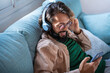 Smiling young man wearing headphones listen to mobile music playing in smartphone app, happy guy relaxing holding using phone enjoy favorite songs relaxing sit on sofa at home. High quality photo