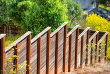 Red Wooden Fence Going Down Hill With Terraced Design And Back And Front Yard Trees In The Midday Sun In The Neighborhood
