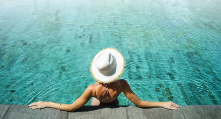 Wall Mural - Back view of young woman wearing summer hat relaxing in big swimming pool with blue water on a sunny day.