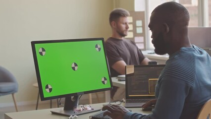African American male software developer typing on keyboard sitting in front of copy space computer monitor, working in modern IT office with colleague