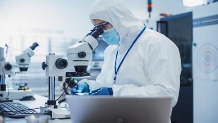 Leinwandbilder - Electronic Manufacturing Factory: Engineer in Sterile Coverall Examining a Circuit Board with Microchips Through a Microscope. Specialist Comparing Results on Laptop Computer