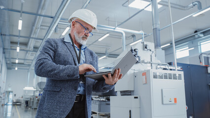 Leinwandbilder - Portrait of a Bearded Middle Aged Engineer Standing in a Factory Facility, Wearing Smart Casual Clothes and a White Hard Hat. Heavy Industry Specialist Working on Laptop Computer.