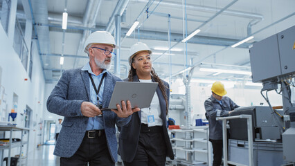 Poster - Two Professional Heavy Industry Engineers Wearing Hard Hats at Factory. Walking and Discussing Industrial Machine Facility, Working on Laptop. African American Manager and Technician at Work.