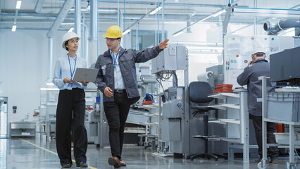 Wall Mural - Two Young Asian Heavy Industry Engineers in Hard Hats Walking with Laptop Computer and Talking in a Factory. Two Manufacturing Employees at Work in Production Facility.