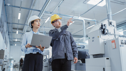 Wall Mural - Two Heavy Industry Employees in Hard Hats Discussing Job Assignments at the Factory, Using Laptop Computer. Asian Engineer and Technician at Work Checking Safety Procedures.