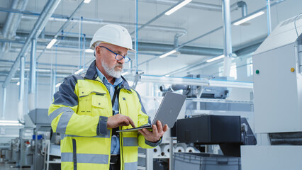 Portrait of a Bearded Middle Aged Engineer Standing in a Factory Facility, Wearing a High Visibility Jacket and a White Hard Hat. Heavy Industry Specialist Working on Laptop Computer.