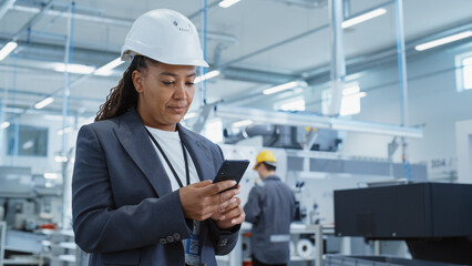 Wall Mural - Portrait of a Black Female Engineer in Hard Hat Standing and Using a Smartphone at Electronics Manufacturing Factory. Technician is Writing a Message and Checking Her Schedule.