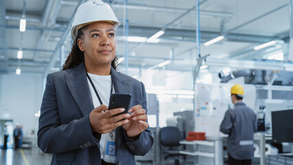 Poster - Portrait of a Black Female Engineer in Hard Hat Standing and Using a Smartphone at Electronics Manufacturing Factory. Technician is Writing a Message and Checking Her Schedule.