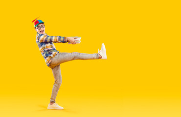 Cheerful preteen boy nerd is funny carrying stack of books on orange background in studio. Schoolboy in funny glasses and cap takes wide step while holding books. Education concept. Full length.