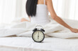 © amorn - Close-up alarm clock on white bed with background woman sitting on bed after waking up in the morning