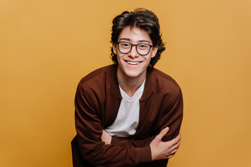 Cheerful caucasian guy in glasses, brown sweatshirt and white t-shirt posing at studio against yellow backdrop toothy smiles. Handsome male teenager with brown hair satisfied indoors. Youth, happiness