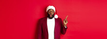 Christmas, Party And Holidays Concept. Impressed Black Man With Beard, Wearing Santa Hat, Pointing Finger Left And Gasping Amazed, Standing Against Red Background