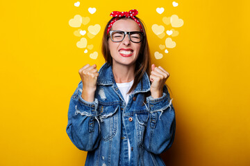 Wall Mural - young woman in a hair band, in a denim jacket clenching her fists on a yellow background, closed her eyes.