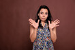 Indian woman showing no problem gesture with palms, looking at camera with confused facial expression. Young lady standing with spread arms portrait, front view studio medium shot on brown background
