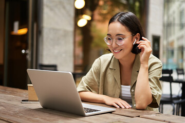 Portrait of asian girl works in cafe, uses laptop and sits outdoors on street. Digital nomad and online learning concept