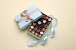 Open box of delicious chocolate candies and light blue ribbon on beige background, flat lay