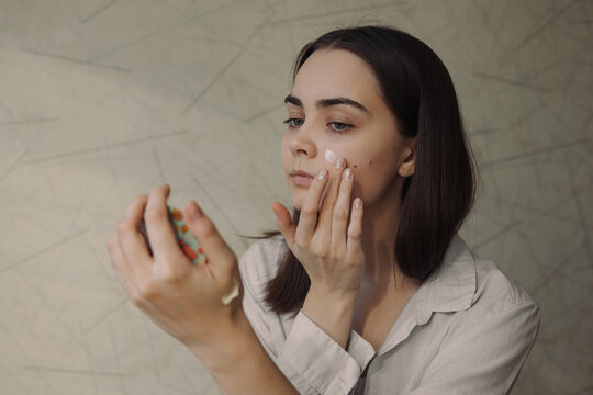 Young beautiful woman applying nourishing face cream and doing daily skin care beauty routine while looking in mirror at home 