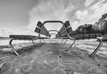 05-11-2021 Riga, Latvia two benches sitting on top of a sandy beach next to a forest of trees and a blue sky with clouds.