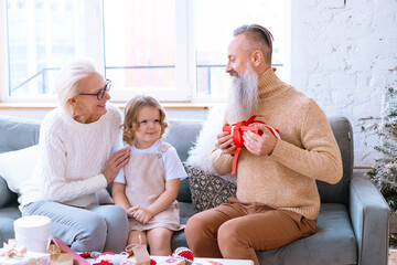 Christmas family grandmother, grandfather and granddaughter sit on sofa near Christmas tree and preparing handmade New Year presents, pretty girl and active happy seniors give each other gift box