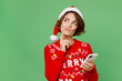© ViDi Studio - Merry young woman wears knitted xmas sweater Santa hat posing hold in hand use mobile cell phone look aside on area isolated on plain pastel light green background Happy New Year 2023 holiday concept