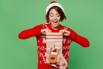 Wall Mural - Merry fun young woman wears knitted xmas sweater Santa hat posing hold in hand look at stocking sock isolated on plain pastel light green background. Happy New Year 2023 celebration holiday concept.