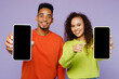 © ViDi Studio - Young couple two friends family man woman of African American ethnicity in casual clothes together hold close up mobile cell phone blank screen look to each other isolated on plain purple background