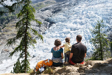 Rear View Of A Family With Toddler Sitting On A Rock And Looking At Bossons Glacier In French Alps. 