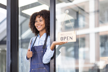 Wall Mural - Happy waitress standing at restaurant entrance. Portrait of african american business woman attend new customers in her coffee shop. Happy woman owner showing open sign in her small business shop.