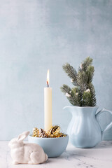 White burning candle, golden decorative cones, white bunnies and branches of Christmas tree in b pitcher on white marble background against blue  textured wall. Rabbit is symbol of 2023.. 