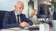 © Bojan - Senior business man wearing formalwear writing on paper in front of computer in office.