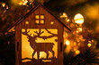 Christmas decoration in the shape of a house with a reindeer inside lit by the warm light of electric garlands on a fir tree. Nice blurred background (bokeh).