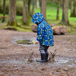 Child wearing a blue rain coat and holding daffodils in his hand, splashing around in a muddy puddle in a park.