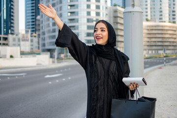 Wall Mural - Young beautiful Arab woman in abaya with shopping bags, phone and wallet hitching a taxi on the street.