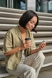 Vertical shot of asian girl drinking her coffee and using mobile phone. Young woman with smartphone and takeaway sits on stairs