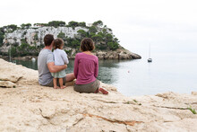 Rear View Of A Family With Toddler Enjoying Views Of Cala Galdana In Menorca From The Edge Of A Cliff On An Autumn Tranquil Day.