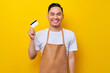 © Bangun Stock Photo - successful smiling young Asian man 20s barista employee wearing brown apron working in coffee shop, holding credit bank card, looking at camera on yellow background. Small business startup concept