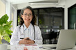 © bongkarn - Cheerful and professional Asian aged female doctor specialist in white gown sitting at her desk