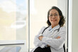 © bongkarn - Successful Asian aged female doctor crossed arms and looking at camera while sitting by the window.