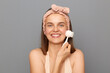 © sementsova321 - Indoor shot of smiling happy woman standing isolated over gray background, using brush for washing, doing morning routine procedures, looking at camera with happy expression, enjoying skin care.