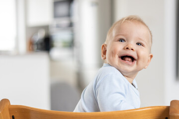 Wall Mural - Happy infant sitting in traditional scandinavian designer wooden high chair and laughing out loud in modern bright home. Cute baby smile