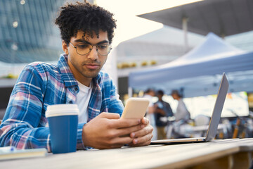 Wall Mural - Curly haired middle eastern man holding mobile phone, reading text message in cafe. Pensive successful freelancer using smartphone, laptop working online sitting at workplace. Technology concept 