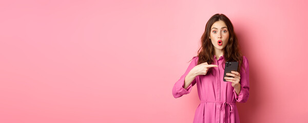Wall Mural - Young woman pointing finger at her phone and gasping wondered, talking about smartphone app, showing something interesting online, standing over pink background