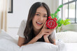 © kaew6566 - Beautiful cheerful asian woman early wake up lying relax on pillow holding hand red alarm clock at home. Happy young girl refreshing early wake up on white with alarm clock looking camera in morning.