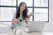 © kaew6566 - Cheerful asian woman playing ukulele sitting on white bed with pillows beside window at apartment. Joyful young girl holding acoustic ukulele in morning at home. Relax female play music ukulele hobby.