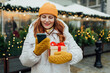 © mdyn - Happy young beautiful woman holding a box gift and sending text message from phone near Christmas decorations, outdoors. Old town