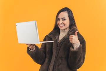 Young adult latin american woman wearing brown fur coat holding up laptop and looking to camera with wide smile and showing thumbs up gesture. Advertising. Online shopping, Yellow background
