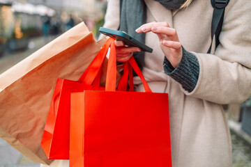 Cropped image of female hands holding red holiday gifts packages used phone on the city street near the shopping center. Retail sale concept. Shopper. Sales. Shopping Center.Cyber Monday.Black Friday