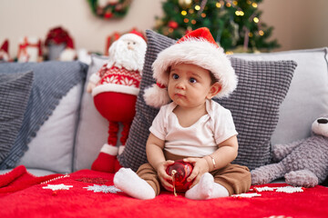 Sticker - Adorable hispanic baby holding christmas decoration ball sitting on sofa at home