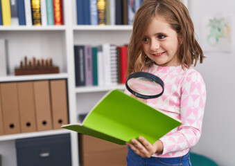 Adorable hispanic girl student reading book using magnifying glass at library school