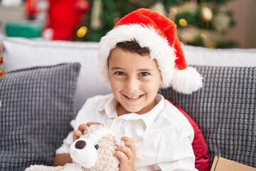 Sticker - Adorable hispanic toddler hugging teddy bear sitting on sofa by christmas tree at home